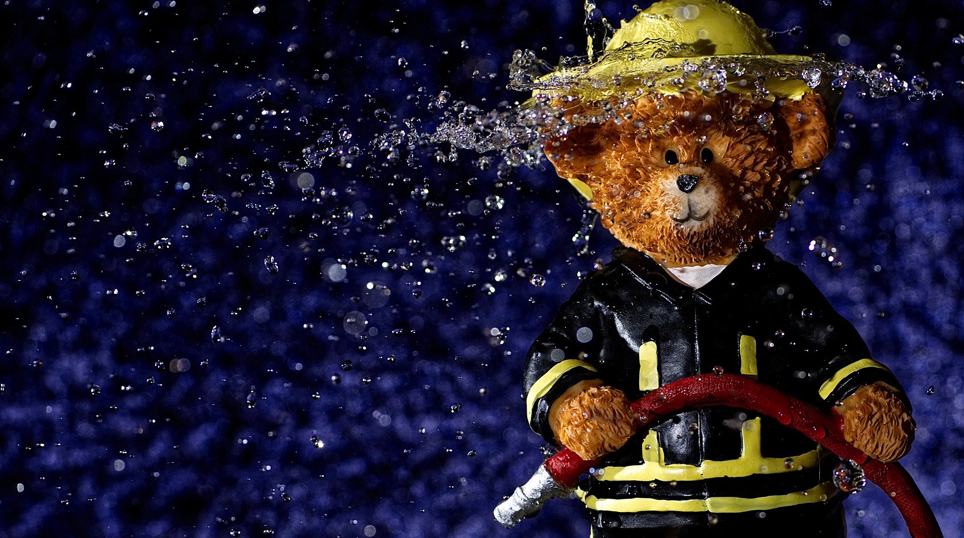 Un ours en peluce porte un costume de pompier. Dans ses mains, une lance à eau l'éclabousse