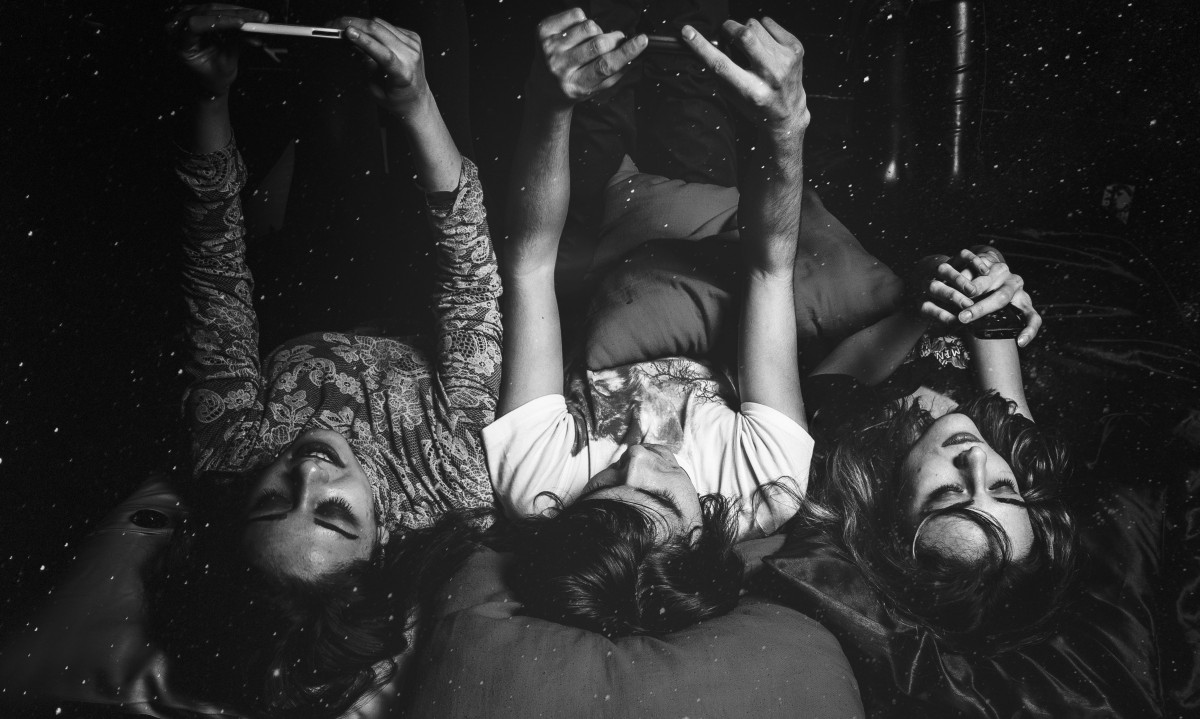 Photo en noir et blanc de trois adolescents couchés sur un lit. Chacun regarde son téléphone portable. 