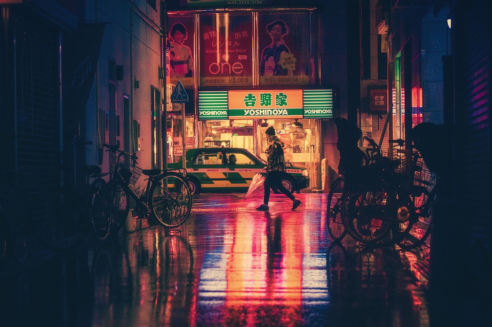 Une photo d'une rue de nuit. La route brille de pluie et les néons flashy éclairent une passante. Des panneaux d'idéogrammes asiatiques emcombrent les façades.